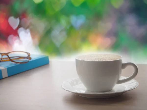 Mesa de oficina de madera con taza de café con leche y gafas modernas en la cubierta azul del libro, hermoso fondo de textura bokeh forma de corazón — Foto de Stock