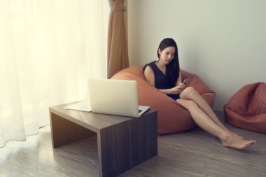 Genç Asyalı kadın portakal fasulye çanta ahşap masa ve beyaz perde windows doku arka plan üzerinde dizüstü bilgisayarınızın arka manzaralı oturma odasında cep telefonu kullanma. ön büro tablosunu görüntülemek.