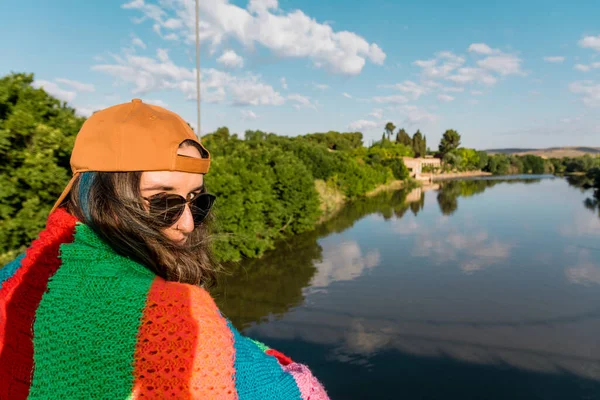 Girl with cap, sunglasses, golden sports jacket and multicolored blanket facing the river at sunrise . Lifestyle concept