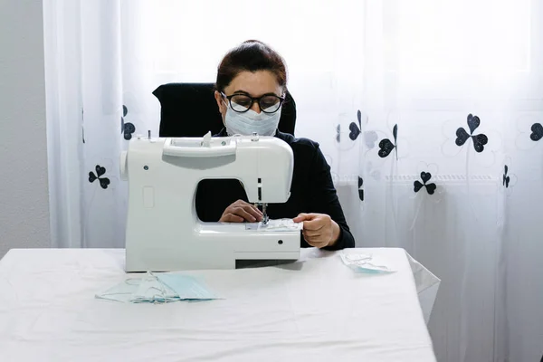 Older woman volunteers sewing masks to combat the virus. Sewing on a white sewing machine. Helping the community. Coronavirus pandemic.