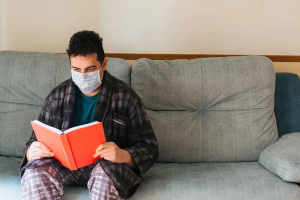 Man in a robe and pajamas sitting on the couch at home with a mask on. Reading a novel. Red book. Concept of quarantine and home confinement. Pandemic coronavirus.