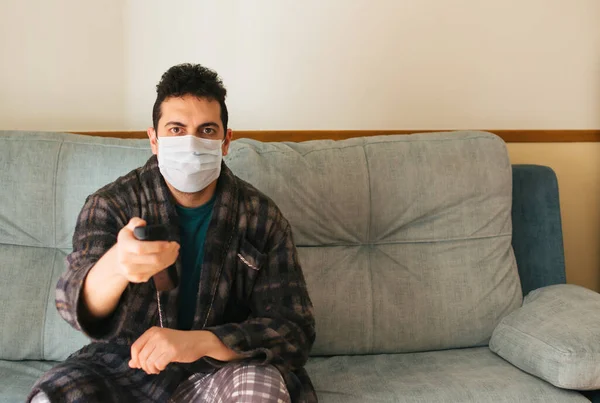 Man in a robe and pajamas sitting on the couch at home with a mask on. Changing the channel with the TV remote. Concept of quarantine and home confinement. Pandemic coronavirus.
