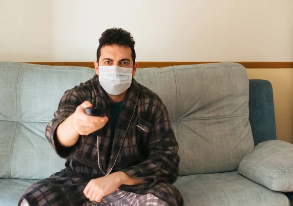 Man in a robe and pajamas sitting on the couch at home with a mask on. Changing the channel with the TV remote. Concept of quarantine and home confinement. Pandemic coronavirus.