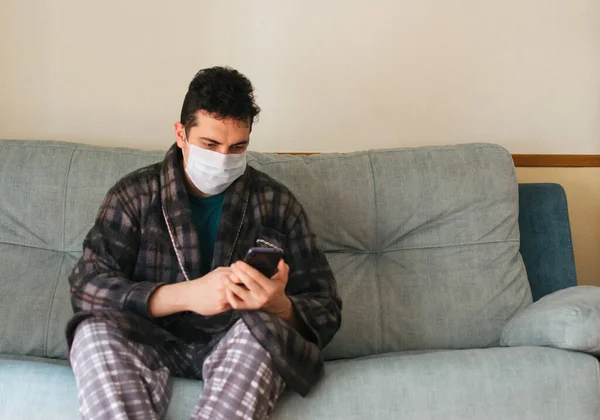 Man in a robe and pajamas sitting on the couch at home with a mask on. Working on his cell phone. Concept of quarantine and home confinement. Pandemic coronavirus.
