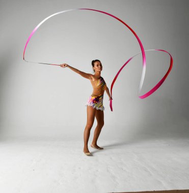 Beyaz arka planda kırmızı kurdeleyle dans eden parlak takım elbiseli bir jimnastikçi.