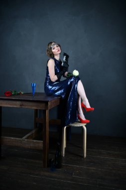 Siyah eldivenli, mavi gece elbisesi giymiş güzel bir kadın elinde silahla bir masada oturuyor.