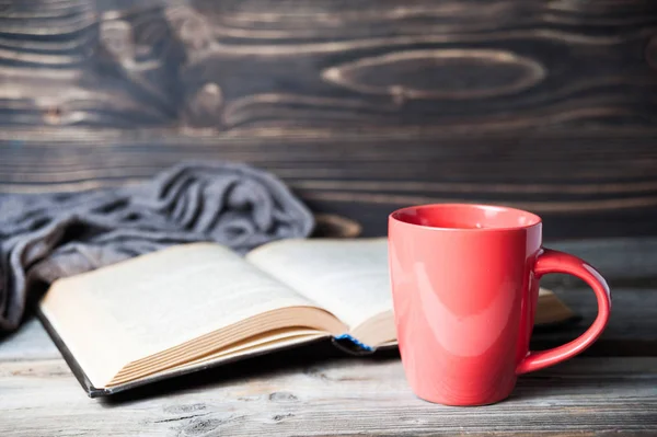 Foto de foco selectivo de gris bufanda de punto acogedor con taza de café o té y libro abierto en una mesa de madera . —  Fotos de Stock