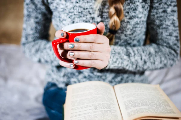 Jeune fille en jeans et pull lit un livre avec une grande tasse de café ou de thé . — Photo