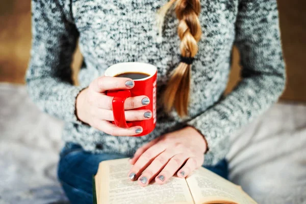 Jeune fille en jeans et pull lit un livre avec une grande tasse de café ou de thé . — Photo