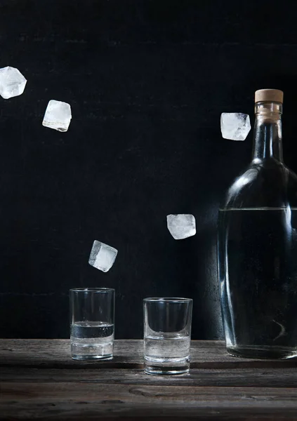 Vodka fria em copos de tiro com gelo levitando voador e garrafa em um fundo preto — Fotografia de Stock
