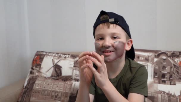 Divertido chico en sombrero con pintura de la cara se sienta en el sofá y incursiones — Vídeos de Stock