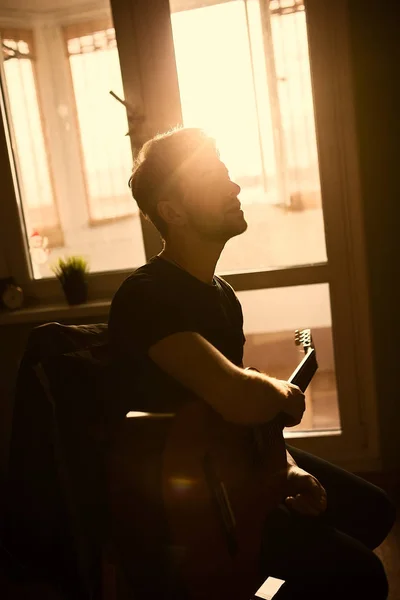 Um tipo atraente a tocar guitarra acústica e a assinar à luz do sol. Loft interior. pessoas estilo de vida . — Fotografia de Stock