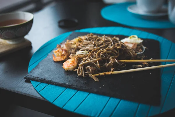 Buckwheat noodles with shrimps and shavings of tuna with sauce and Chinese sticks on black plate. Asian food background. Eating concept. Restaraunt place with wooden table. Flare copy space for text,