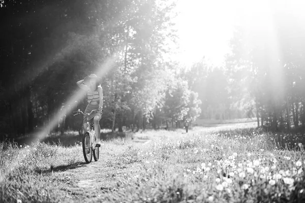 Funny girl driving bicycle outdoor. Sunny summer lifestyle concept. Woman in dress and hat in Field with dandelions. Female ride in park. Light effect for text. Copyspace for design. BW photo. — Stock Photo, Image
