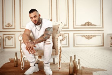 Moda dövme ve otel iç altın dekor ile beyaz rahat kıyafetler sandalyede oturan bir siyah sakallı şık yakışıklı bir erkek model seksi closeup portresi. Profesyonel Studio resim