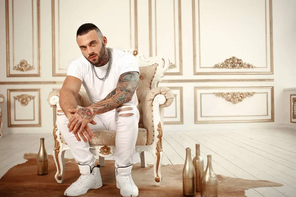 Moda dövme ve otel iç altın dekor ile beyaz rahat kıyafetler sandalyede oturan bir siyah sakallı şık yakışıklı bir erkek model seksi closeup portresi. Profesyonel Studio resim
