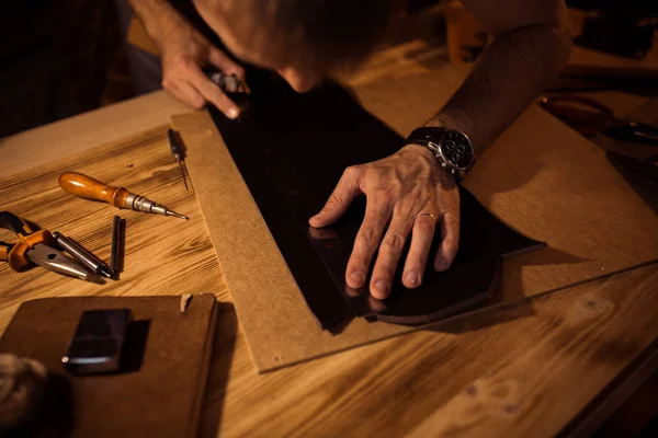 Arbeitsprozess des Ledergürtels in der Lederwerkstatt. Mann hält Werkzeug in der Hand und arbeitet. Gerber in der alten Gerberei. Holztisch Hintergrund — Stockfoto