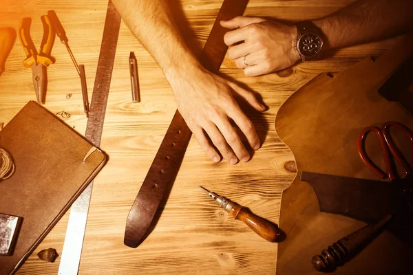 Processo di lavorazione della cintura in pelle nell'officina. Uomo che si tiene per mano su un tavolo di legno. Strumenti di creazione su sfondo. Abbronzatura nella vecchia conceria. Chiudete il braccio agli uomini. Luce calda per testo e design — Foto Stock