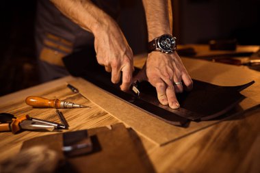 Working process of the leather belt in the leather workshop. Man holding crafting tool and working. Tanner in old tannery. Wooden table background clipart