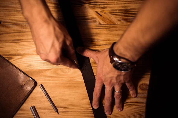 Arbeitsprozess des Ledergürtels in der Lederwerkstatt. Mann hält Werkzeug in der Hand und arbeitet. Gerber in der alten Gerberei. Holztischhintergrund. Nahaufnahme Mann Arm — Stockfoto