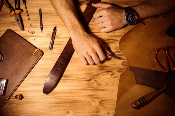 Processo di lavorazione della cintura in pelle nell'officina. Uomo che si tiene per mano su un tavolo di legno. Strumenti di creazione su sfondo. Abbronzatura nella vecchia conceria. Chiudi braccio uomini — Foto Stock