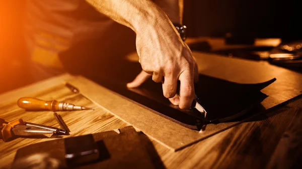 Proceso de trabajo del cinturón de cuero en el taller de cuero. Hombre sosteniendo herramienta. Tanner en curtiduría vieja. Fondo de tabla de madera. Cierra el brazo del hombre. Luz cálida para texto y diseño. Tamaño del banner web — Foto de Stock