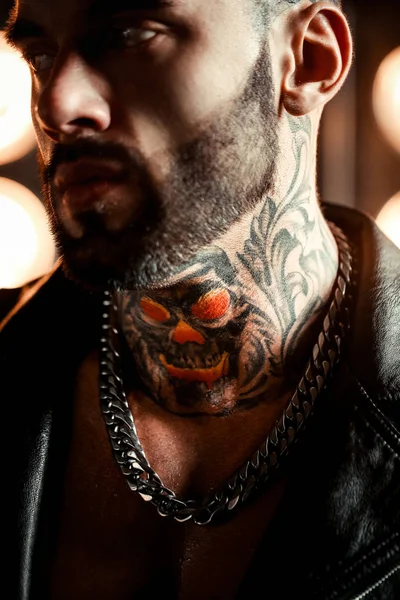 Sexy closeup portrait of brutal handsome male model in fashion leather jacket and with a black beard. Tattoo skull and eye. Strong man. Background with vintage retro lights.