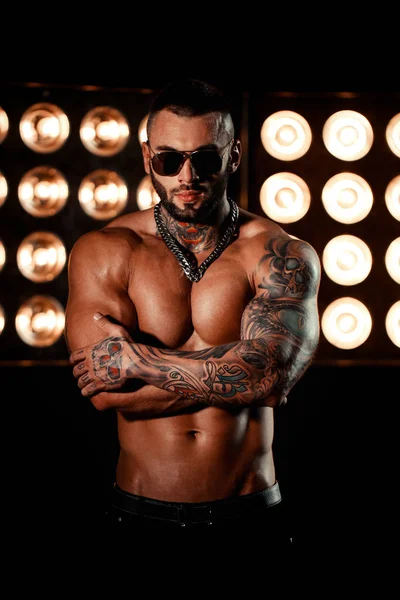 Sexy closeup portrait of brutal handsome male model with a black beard and in sunglasses. Tattoo skull and eye. Strong man with abs and tan. Background with vintage retro lights.