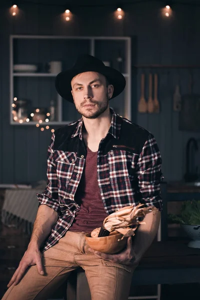 Un homme dans la cuisine. Photo de style de vie couleur rustique. Les gens et une saine alimentation concept. Portrait de gars souriant en chapeau noir avec des légumes . — Photo