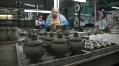 Çanak çömlek, sürahi ve semaverler, en üstte mini çaydanlıklar var. Arka planda iki kadın birbiri ardına oturup çömlekçilikle çalışıyor. Sağdan sola kolayca dolambaçlı yol ile Panorama.