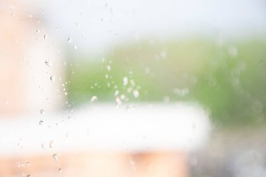 Arka planda, pencereden turuncu bir binaya ve ağaçlara odaklanmış, camda yağmur damlaları var. Üzücü, yağmurlu bir gün.