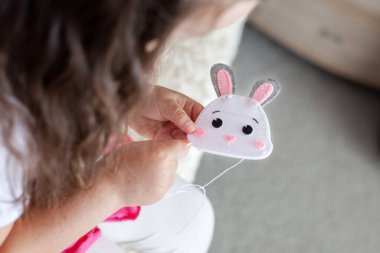 Çocuk kız bej bir battaniyenin üzerinde keçeden yapılmış bir oyuncak dikiyor; dikiş işi.