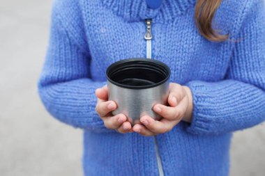 Mavi örülü süveter giyen genç bir kız termostan çay ya da kahve içer.