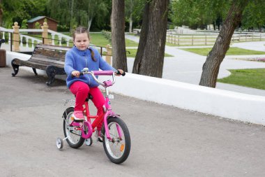 Mavi kazaklı ve pembe pantolonlu bir kız nehir kıyısında pembe bir bisiklet sürüyor.