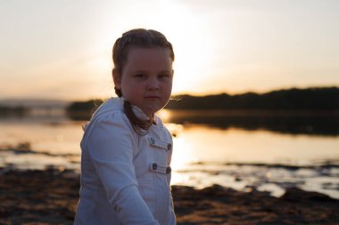 Bir kız gün batımında nehir kıyısında kumlu bir plajda kumla oynuyor.