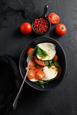 Kiraz domatesleri, mozzarella bufalo peyniri fesleğen, pesto sosu ve siyah tahtadaki baharatlar. İtalyan geleneksel Caprese salata malzemeleri. Siyah kasede çatallı Akdeniz yemeği.