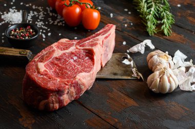 Koyu ahşap masa arkasında çiğ Kovboy bifteği. Ribeye Bifteği, Bone-In bifteği veya baharatlı BBBQ bifteği, biberiye, domates, chikie, sarımsak, biber, biber, et bıçağı ile tuz.