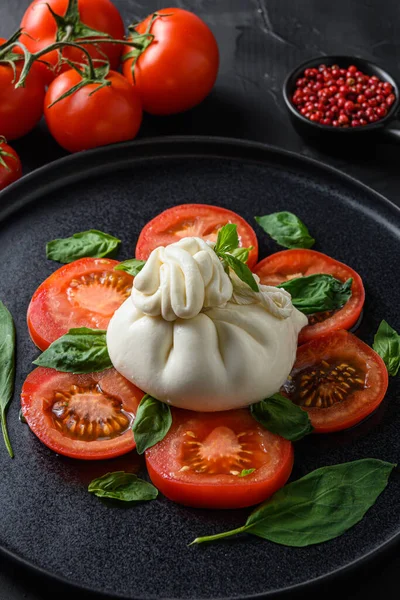 Bufalo burrata peyniri taze domates ve fesleğen yapraklarıyla servis edilir. Siyah bir taş arka planda.