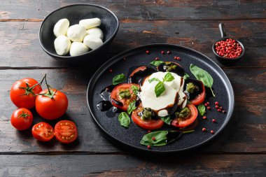 İtalyan yemeği - dilimlenmiş domatesli caprese salatası, mozzarella peyniri, fesleğen, zeytinyağı. Koyu ahşap arka planda siyah tabakta servis edilmiş. Üst Manzara. Kırsal biçim.