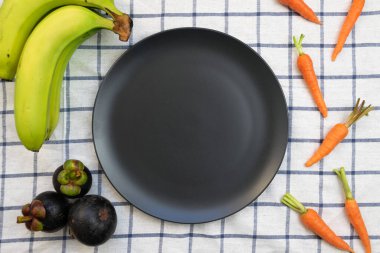 Dümdüz uzanmış, bebek havuçlarla çevrili boş siyah tabak, yeşil muz ve mavi örgülü beyaz masa örtüsü üzerine mangosteen. Sağlıklı yaşam.