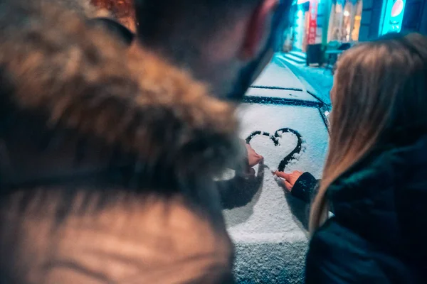 Young adult couple draws a heart on snow covered car — ストック写真