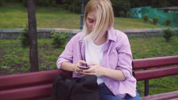 Akıllı telefon kullanan ve bankta oturan genç bir kız. — Stok video