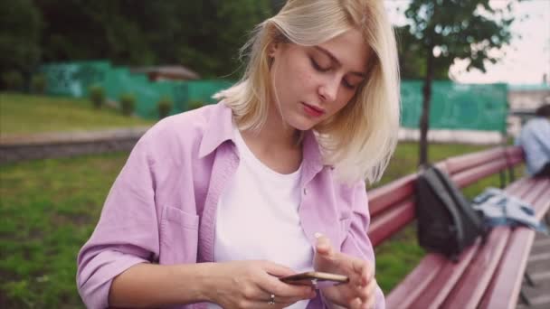 Adolescente chica usando un teléfono inteligente y mensajes de texto sentado en un banco — Vídeo de stock