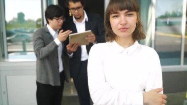 Kameraya poz veren genç ve güzel bir kadın. Arka planda iki iş adamı.