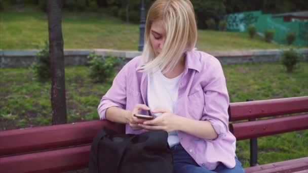 Adolescente chica usando un teléfono inteligente y mensajes de texto sentado en un banco — Vídeo de stock