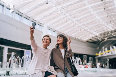 İki kız alışveriş merkezinde selfie çekiyor, arka planda bir çeşme.