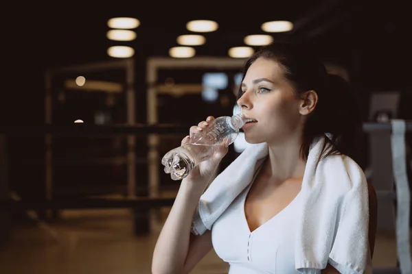 Прекрасная молодая женщина с полотенцем на плечах пьет воду из бутылки в спортзале — стоковое фото