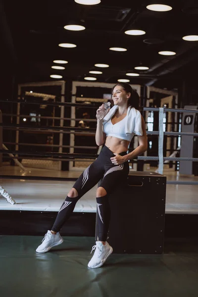 Hermosa joven con una toalla en los hombros bebiendo agua de una botella en el gimnasio — Foto de Stock