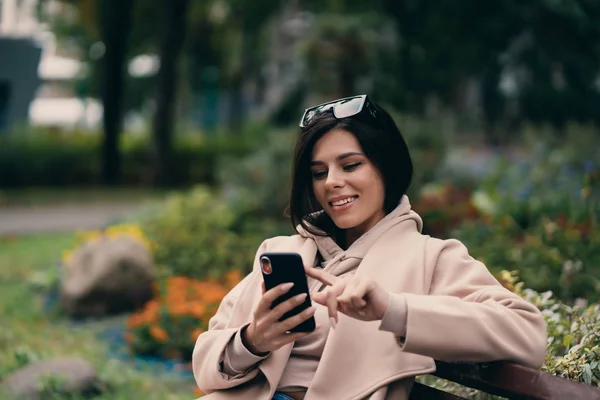 Menina feliz usando um telefone inteligente em um parque da cidade sentado em um banco — Fotografia de Stock