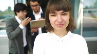 Kameraya poz veren genç ve güzel bir kadın. Arka planda iki iş adamı.